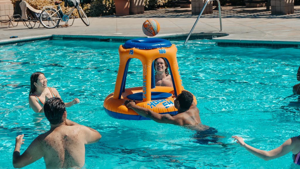 A group of friends having fun playing basketball with an inflatable hoop in a swimming pool during summer.
